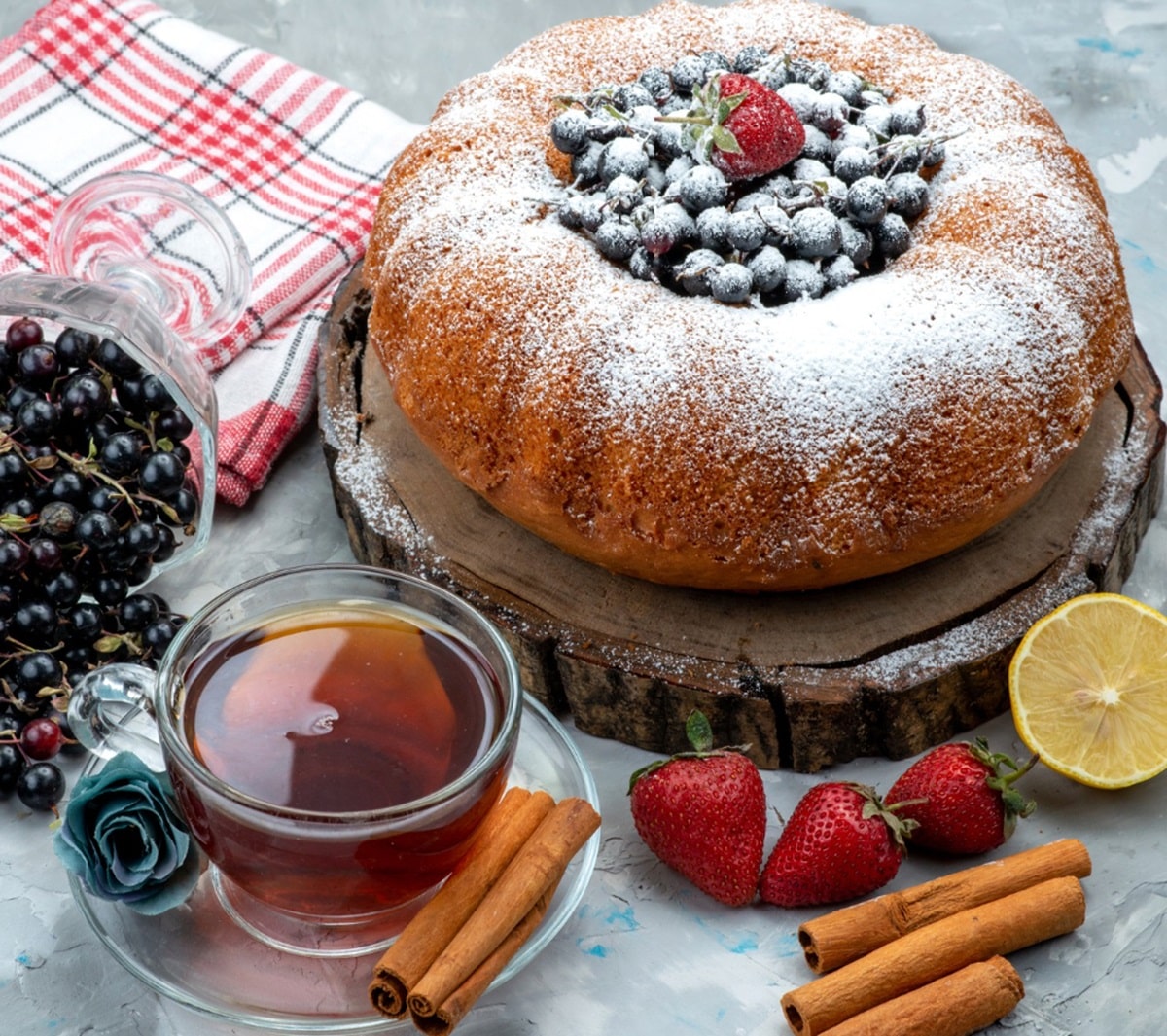 Bolo caseiro decorado com açúcar de confeiteiro e frutas vermelhas no topo, acompanhado de uma xícara de chá, paus de canela, morangos e groselhas, sobre uma tábua rústica de madeira.