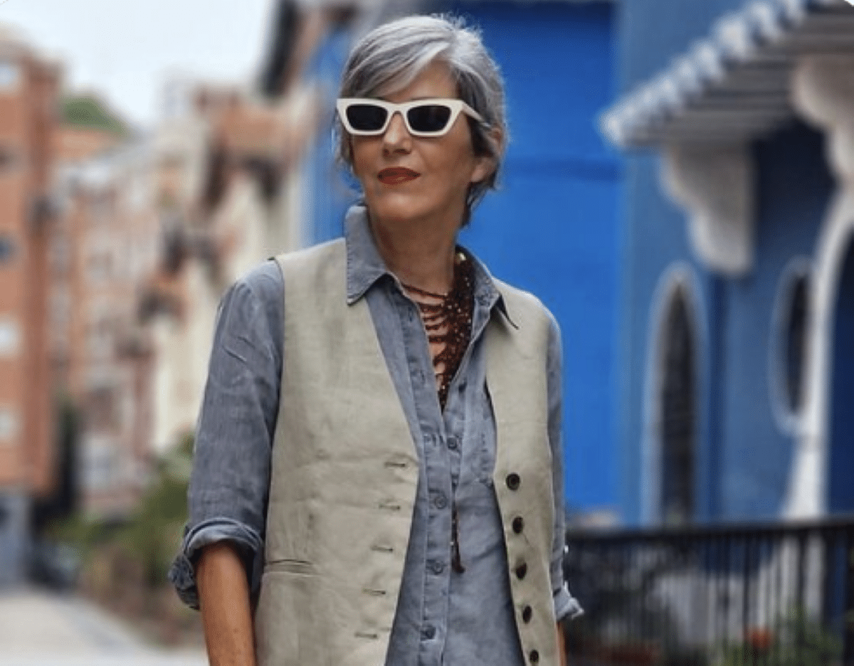 Mulher idosa com cabelo grisalho e óculos de sol brancos, usando colete bege sobre camisa cinza, em uma rua colorida.