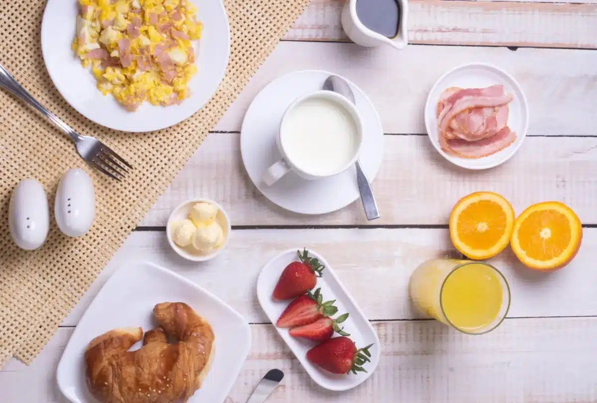 Café da manhã nutritivo com croissant, frutas, ovos mexidos, bacon, leite e suco de laranja.