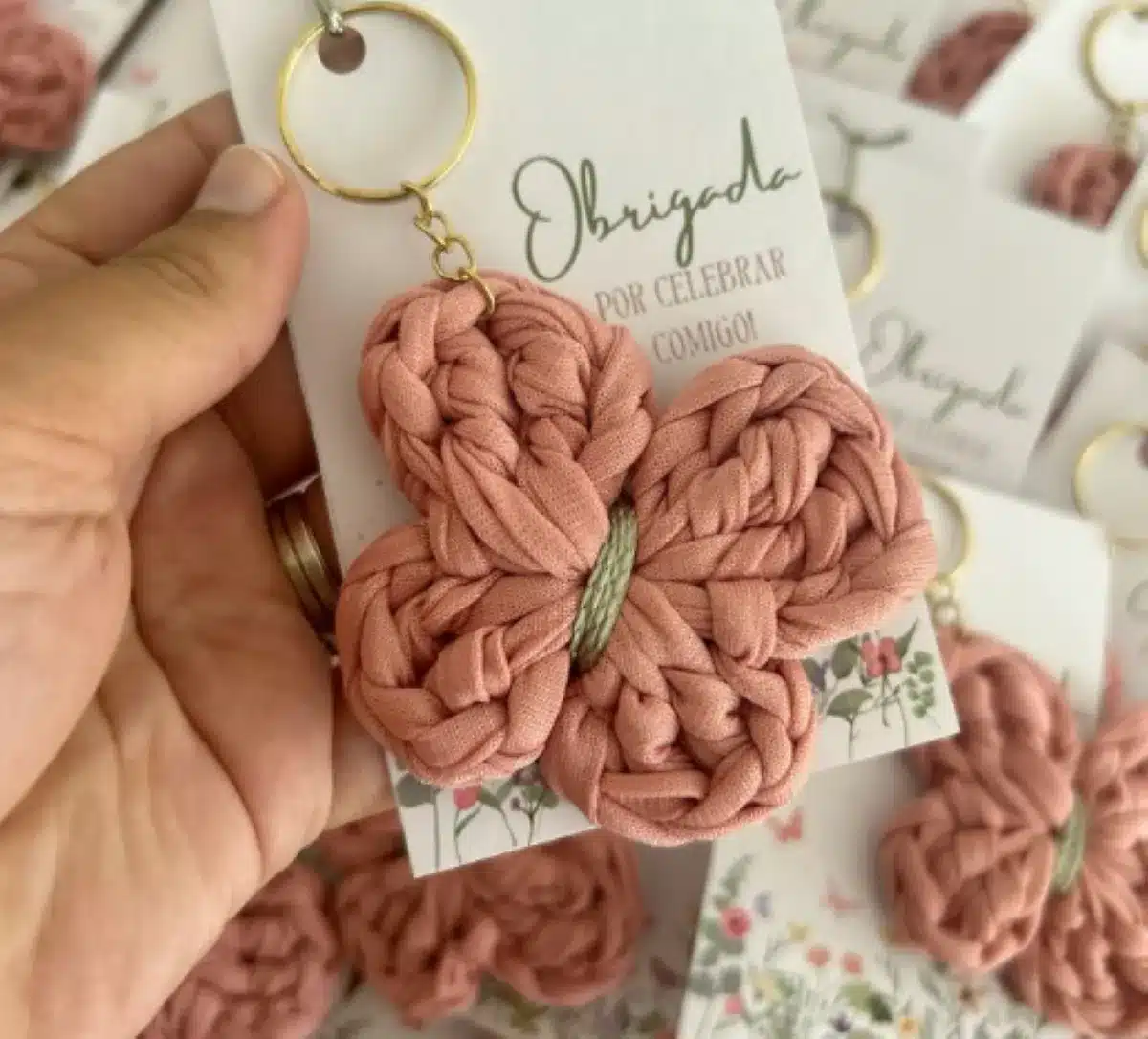 Chaveiro de crochê em forma de borboleta rosa, com detalhes em verde, preso a uma chaveiro dourado.