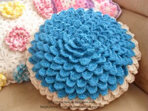 Almofada de crochê redonda com efeito escama de dragão em azul e bege, trazendo um visual sofisticado e artesanal.