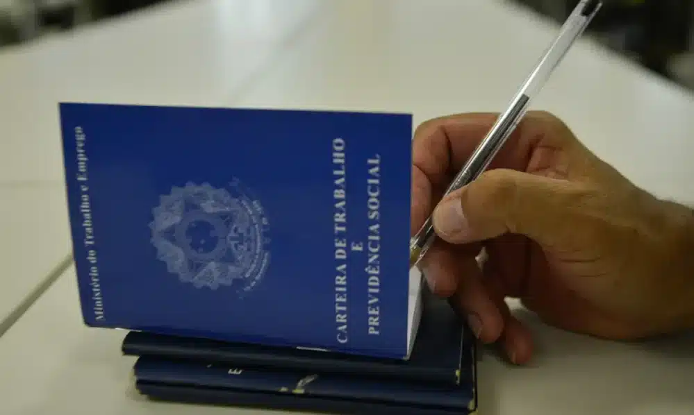 Pessoa assinando carteira de trabalho azul do Ministério do Trabalho e Previdência Social.