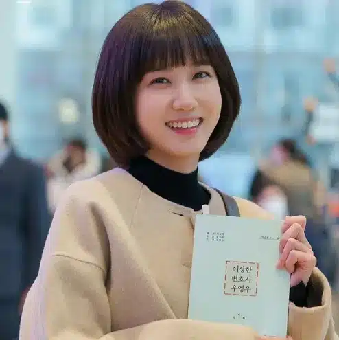 Mulher sorrindo e segurando um livro, vestindo um casaco bege.