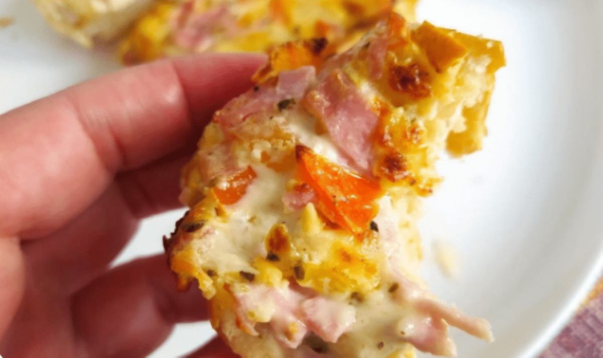 Pedaço de pão salgado de presunto e queijo com pedaços de tomate e crosta dourada, segurado à mão
