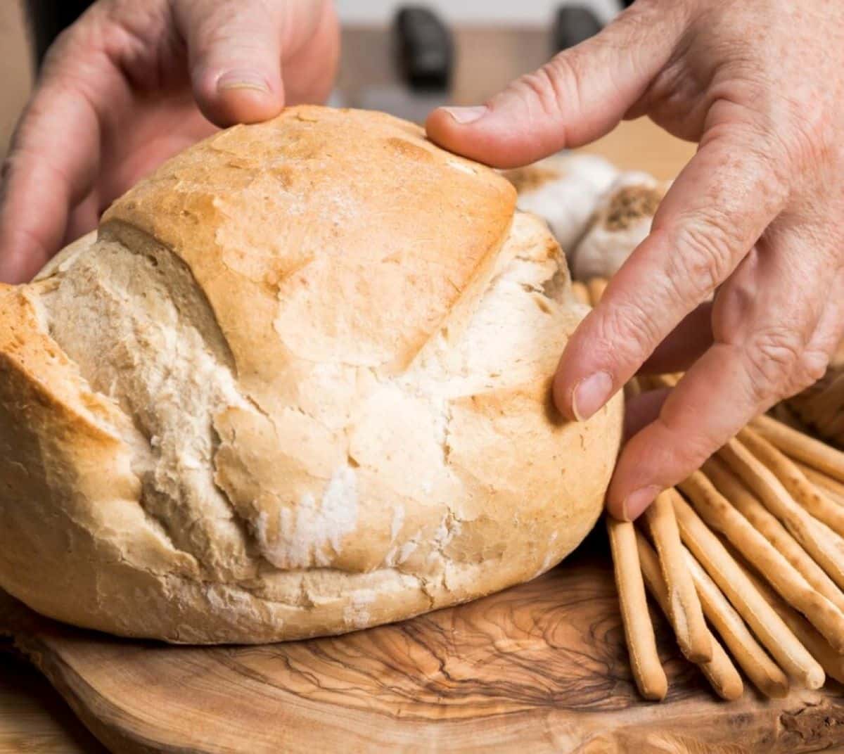 Mãos segurando pão caseiro fresco em tábua de madeira, ao lado de grissinis.