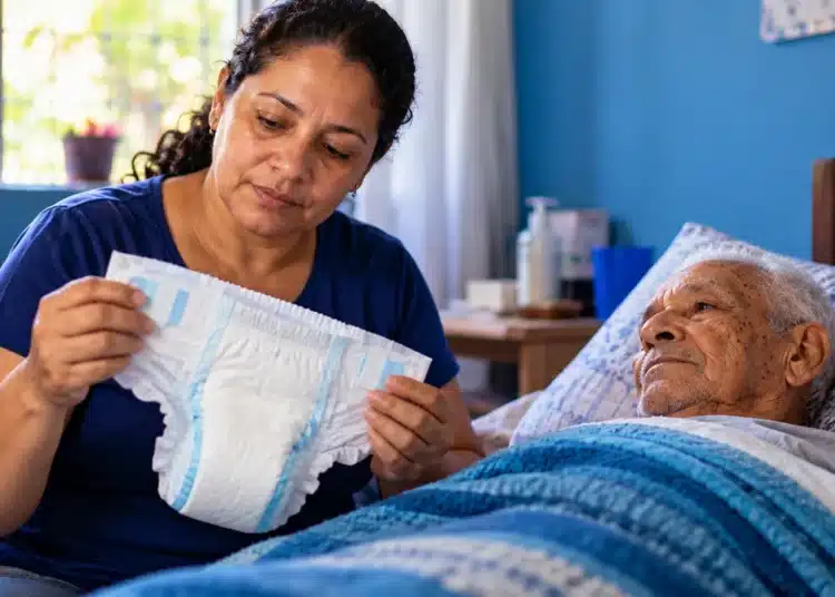 Cuidadora segurando fralda geriátrica ao lado de idoso acamado.