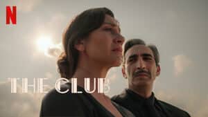 Mulher com expressão séria olhando para o horizonte, acompanhada por um homem de terno, cena da série turca 'The Club' na Netflix.