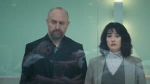 Homem e mulher com expressões sérias observam através de um vidro, cena da série turca 'Amantes Anônimos'.