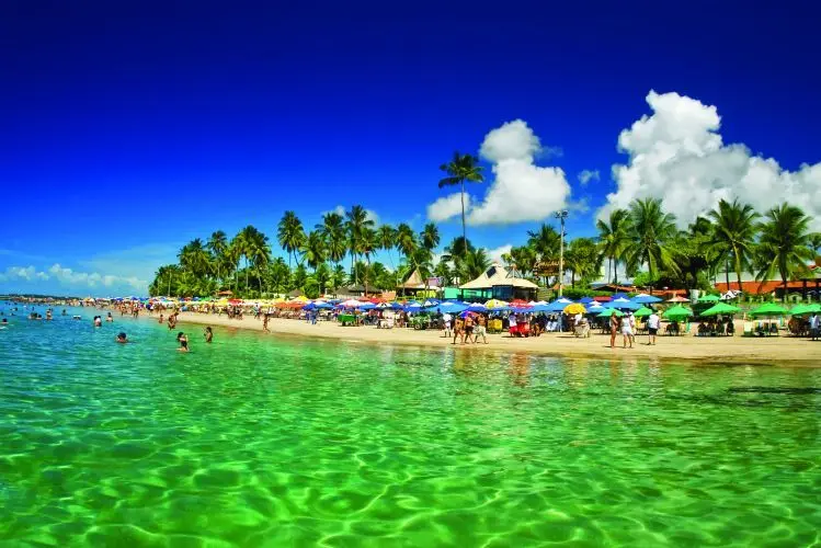 Beleza e tranquilidade em Porto de Galinhas Vista de Porto de Galinhas, com águas cristalinas, coqueiros e barracas coloridas à beira-mar.