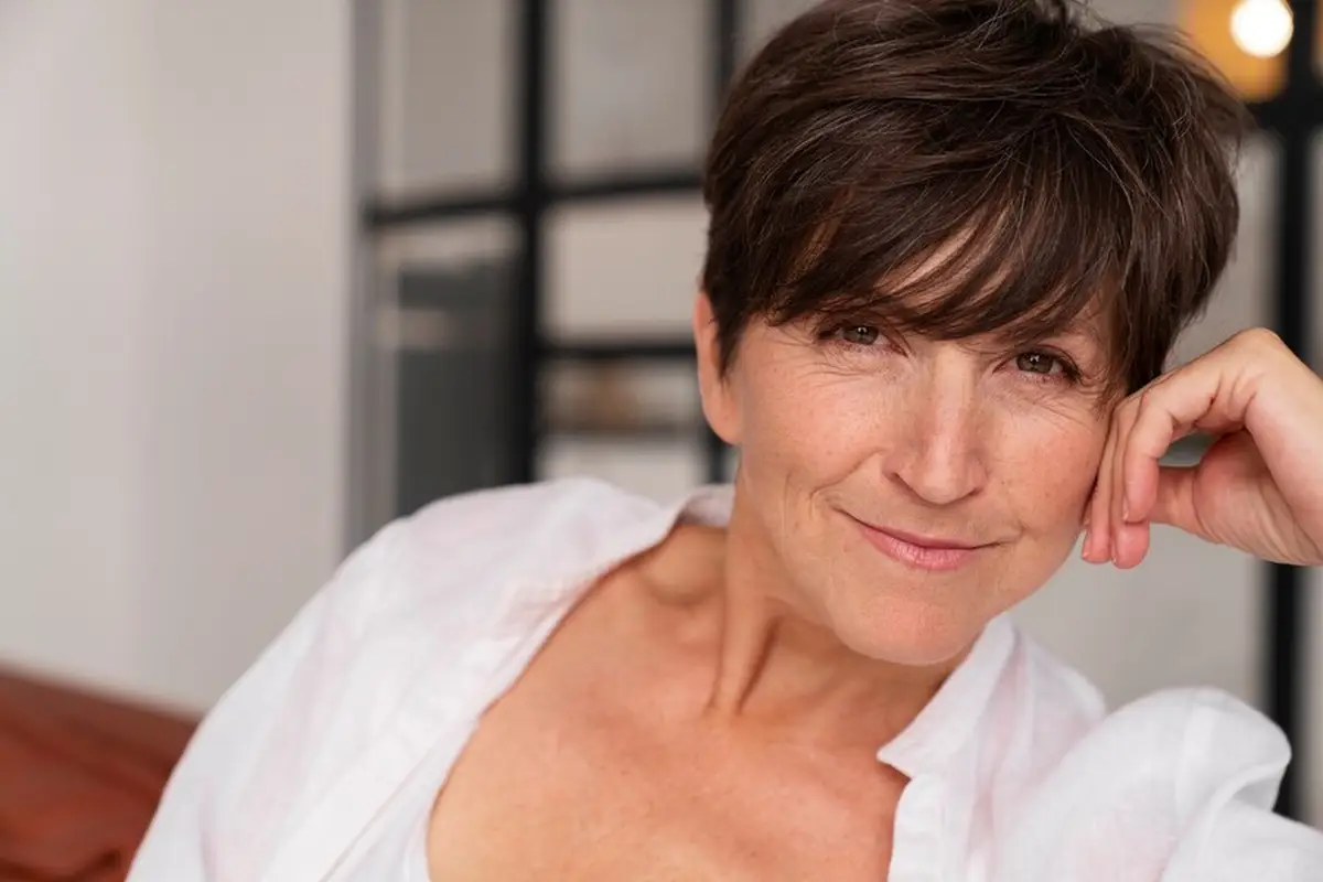 Mulher com cabelo curto e castanho escuro, sorrindo suavemente, vestindo uma blusa branca, representando a tendência de cores para cabelo no inverno de 2025.