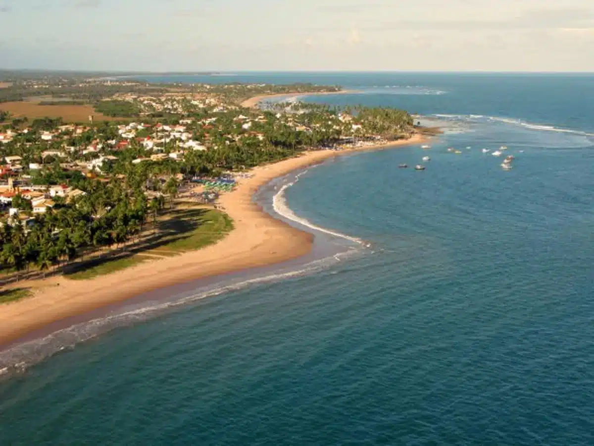 Vista aérea da praia de Guarajuba em Camaçari, Bahia, com águas claras e áreas de vegetação ao fundo.