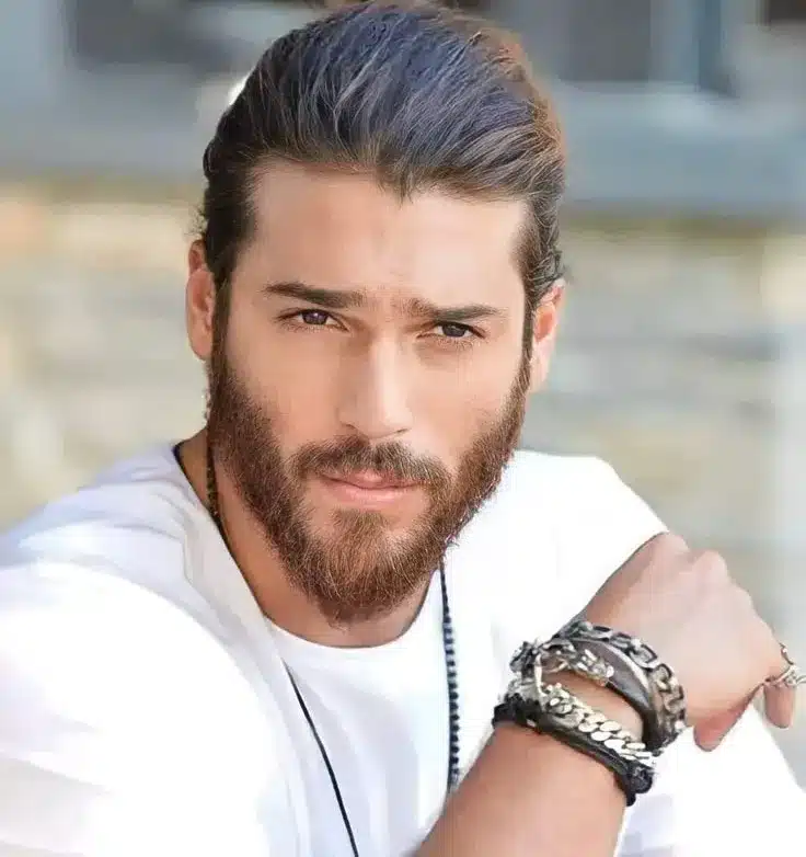 Can Yaman, ator turco com cabelo preso e barba, usando uma camiseta branca e acessórios, olhando para a câmera.