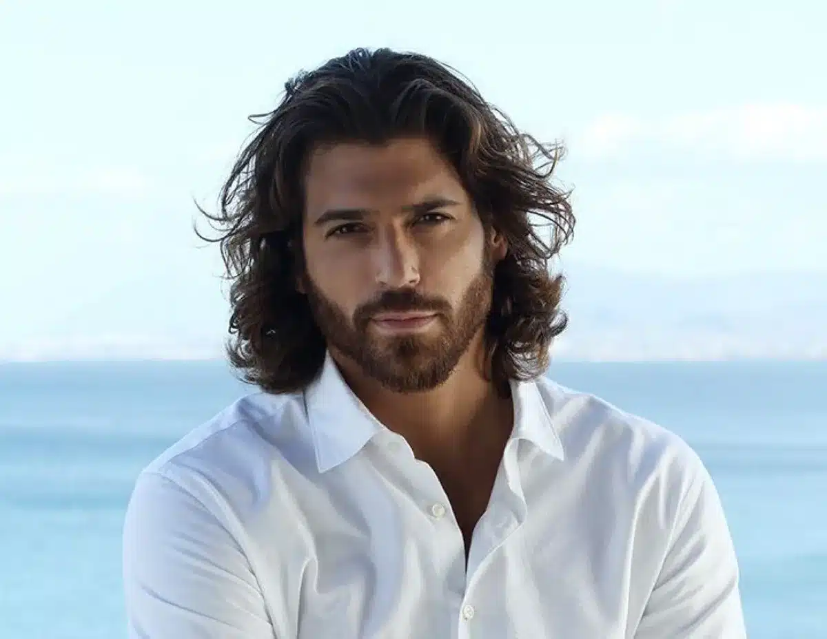 Can Yaman, ator turco com cabelo ondulado e barba, posando com camisa branca ao fundo do mar.