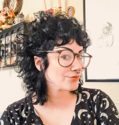  "Mulher com cabelo cacheado no estilo mullet, usando óculos de grau e um visual moderno e descontraído."