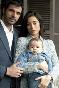 Imagem de uma cena da novela turca 'Sıla: Prisioneira do Amor', mostrando um casal com seu filho. O homem e a mulher estão vestidos de forma elegante, com a mulher segurando o bebê. Eles estão em um ambiente acolhedor e familiar.