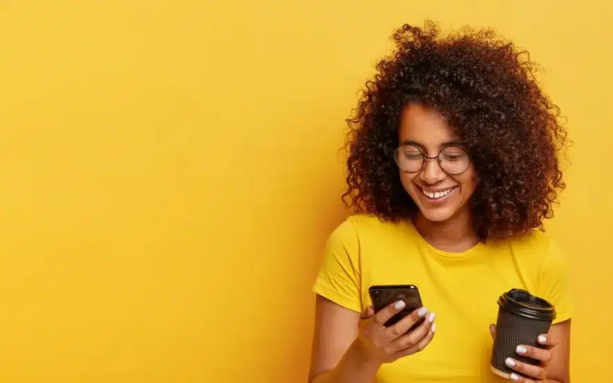 Mulher sorridente segurando um celular e um copo de café contra um fundo amarelo.