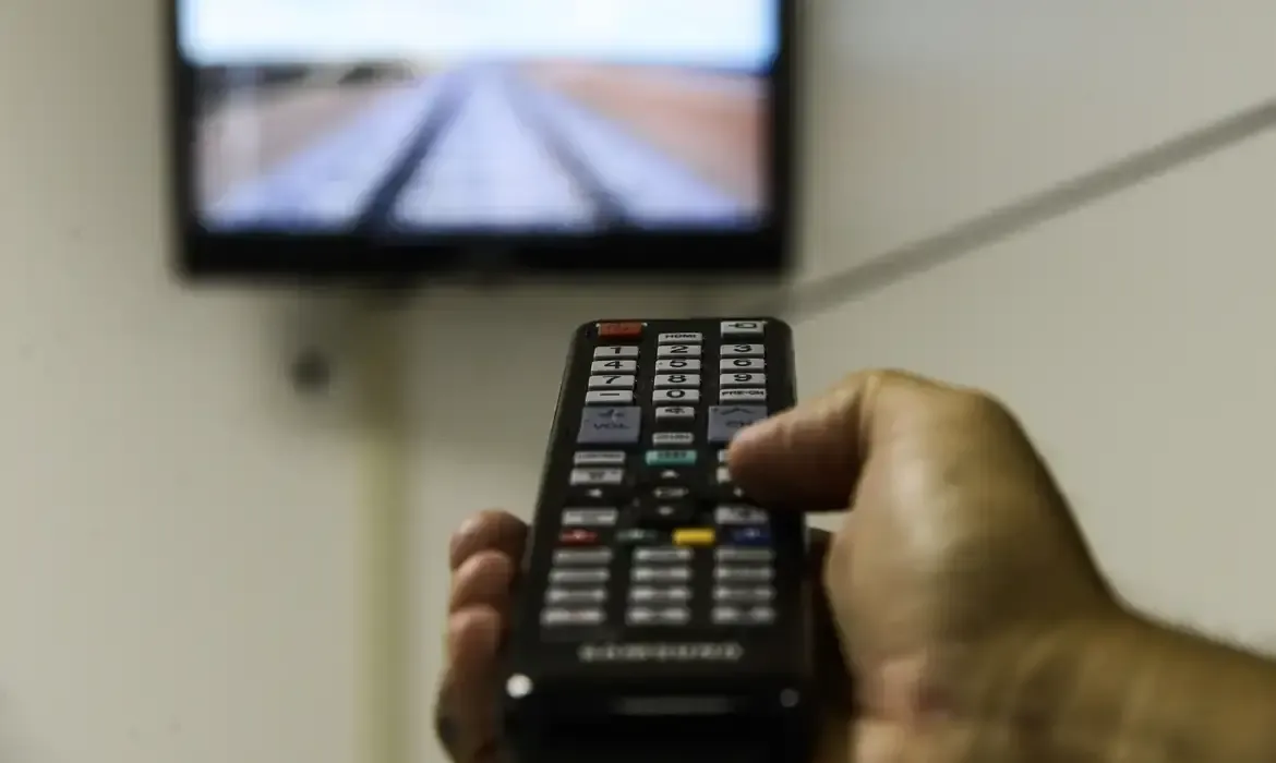 Pessoa segurando controle remoto de TV, enquanto assiste à tela, ilustrando a troca gratuita da antena parabólica digital.