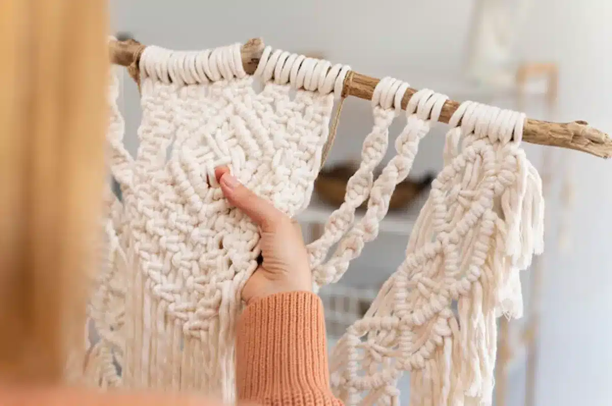 Pessoa segurando um pedaço de macramê pendurado em um galho, com fios brancos e um design detalhado.