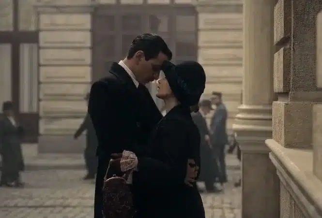 Imagem da série 'Meia-Noite no Hotel Pera Palace' com um casal se abraçando e se olhando com carinho em um cenário vintage, ambientado em um cenário urbano clássico.