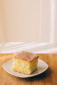 Fatia de bolo de fubá com maizena e erva-doce servida em um prato branco sobre uma tábua de madeira, com açúcar cristal polvilhado por cima.