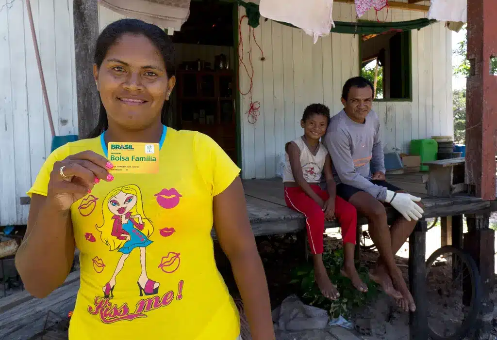 Mulher segurando o cartão do Bolsa Família com a família ao fundo, mostrando o apoio do programa.
