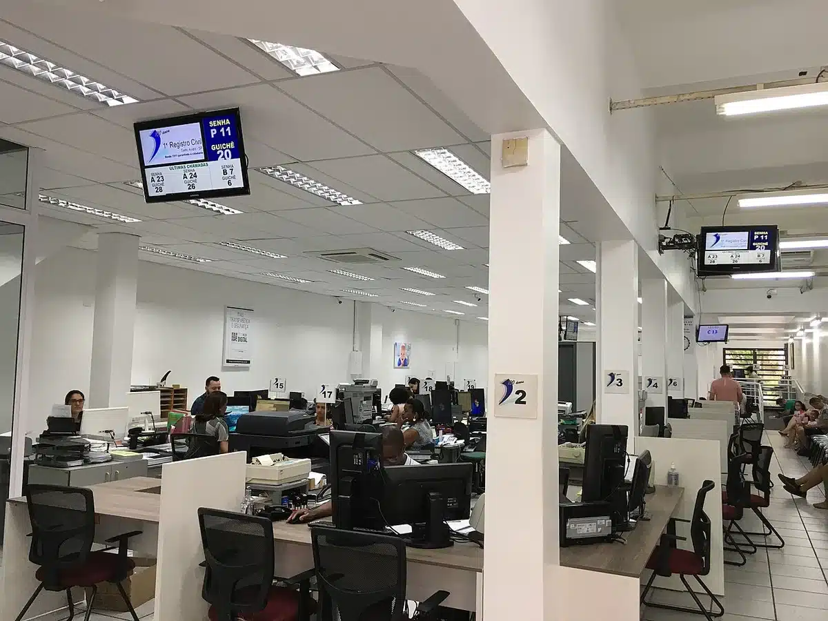 Ambiente interno de um cartório com funcionários atendendo ao público e telas exibindo informações de senhas.