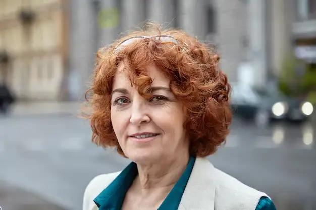 Mulher com cabelo ruivo acobreado, com um sorriso gentil, usando uma camisa azul e um casaco claro, destacando a tonalidade vibrante e cheia de personalidade do ruivo acobreado.