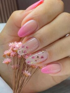 Unhas alongadas em gel com acabamento rosa e decoração floral delicada, segurando flores secas.