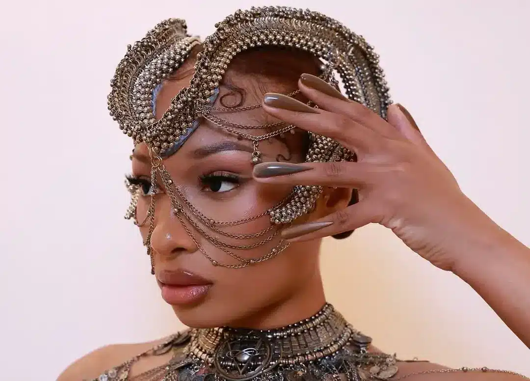 Megan Thee Stallion com estilo de unhas invertidas, usando um look de alta-costura metálico e detalhado, exibindo um visual audacioso e sofisticado.