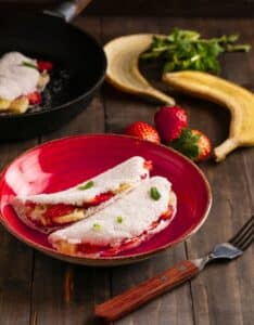 Tapioca recheada com banana e morango em prato vermelho sobre mesa de madeira