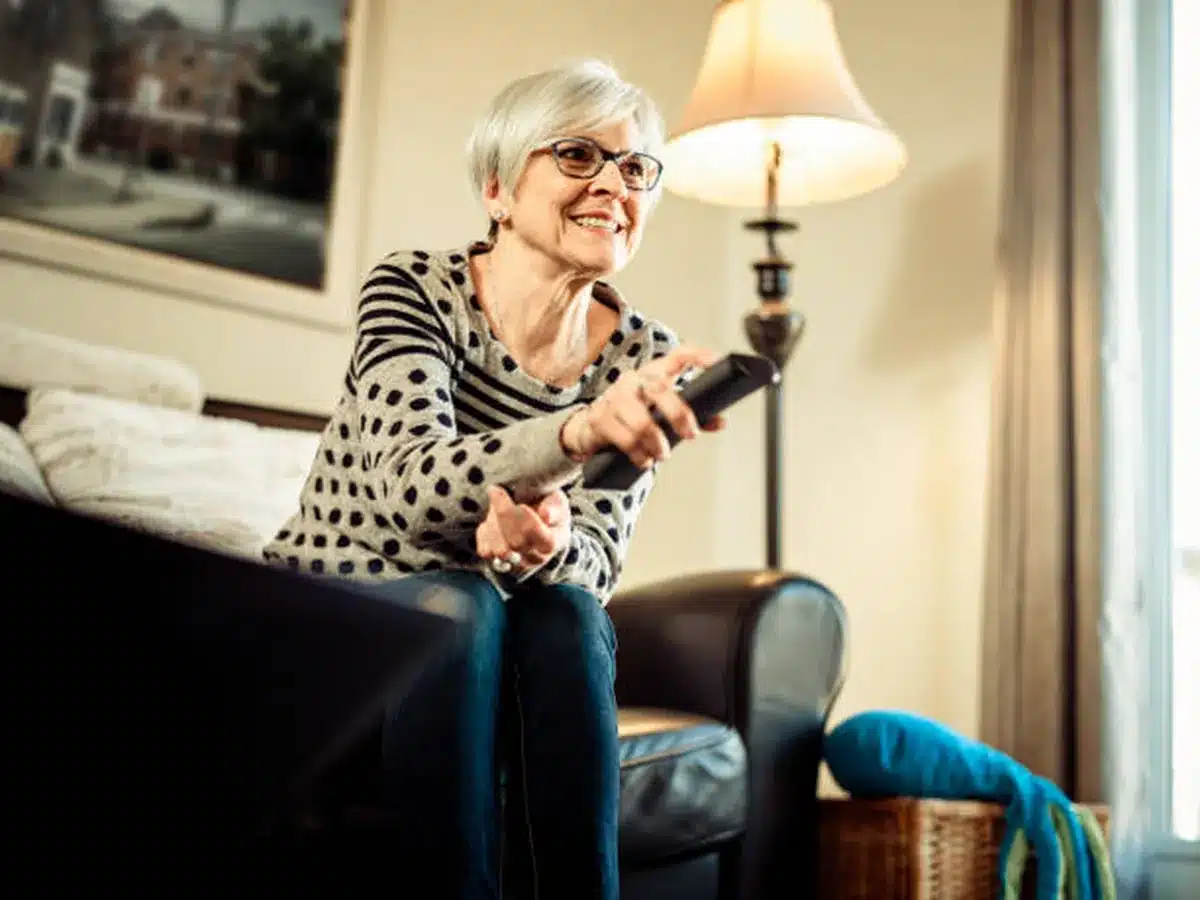 Senhora sorridente assistindo TV com controle remoto em mãos, em uma sala aconchegante.