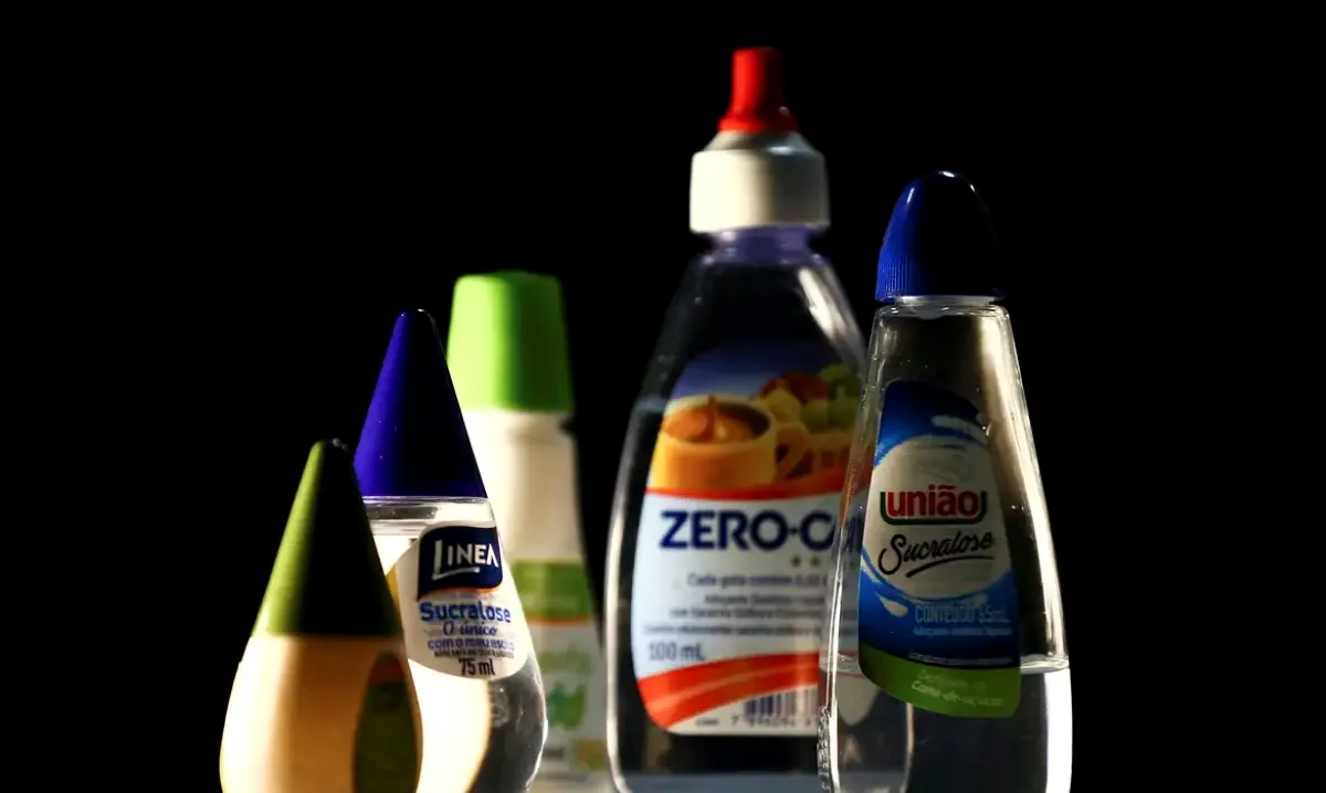 Diversos frascos de adoçantes líquidos de marcas diferentes sobre fundo escuro.