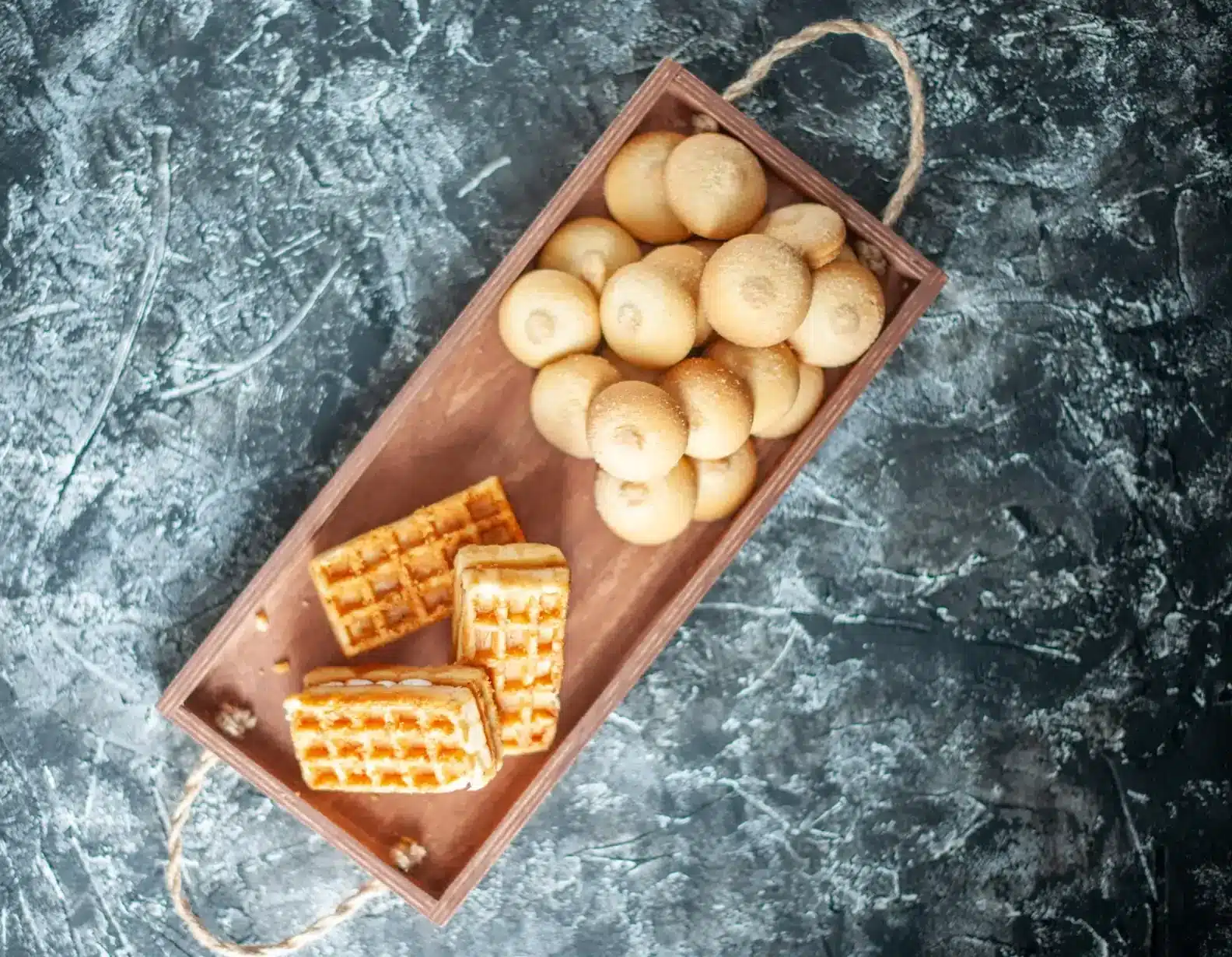 Waffles de pão de queijo e pãezinhos servidos em bandeja de madeira sobre fundo escuro.