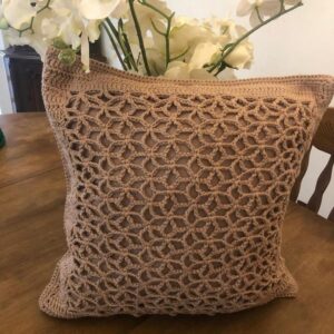 Almofada de crochê na cor Mocha Mousse com padrão delicado de pontos e flores brancas ao fundo.