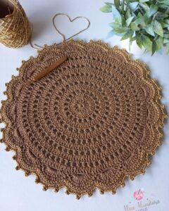 Sousplat de crochê na cor Mocha Mousse com detalhes dourados, fio e agulha ao fundo.