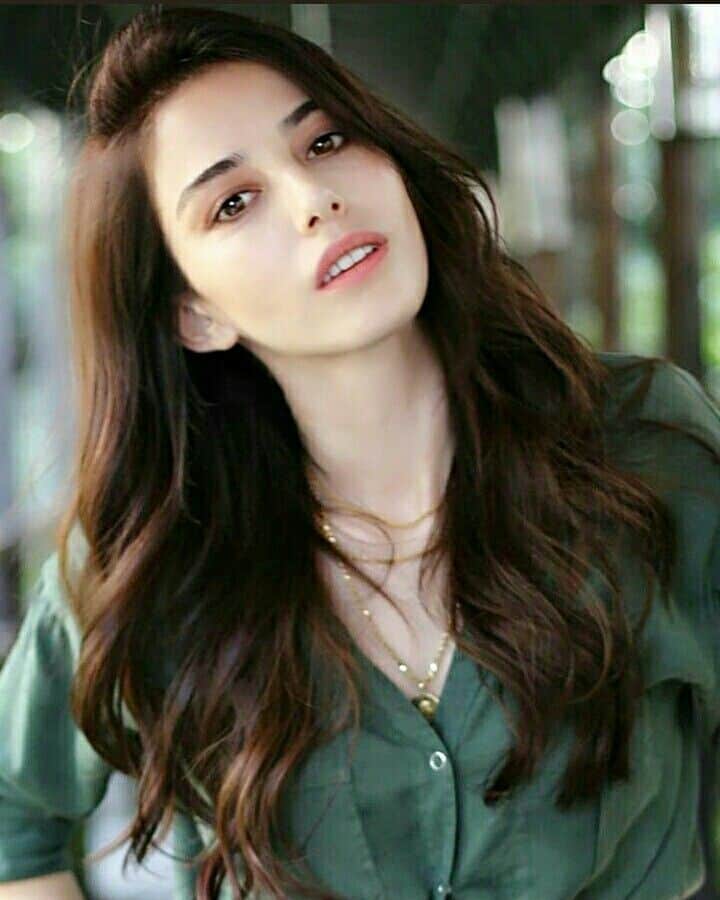 Atriz turca Özge Gürel com cabelos longos e ondulados, vestindo blusa verde, em ambiente com luz natural.