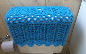 Capa azul de crochê cobrindo caixa de descarga no banheiro com acabamento em ponto alto vazado.
