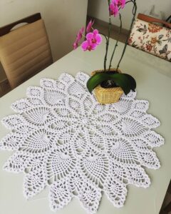 Centro de mesa redondo em crochê branco com vaso de orquídeas sobre mesa de jantar clara