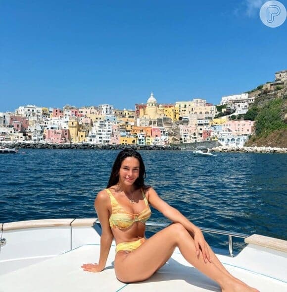 Atriz turca Ebru Şahin sorrindo, sentada na proa de um barco, com fundo de cidade costeira e mar azul.