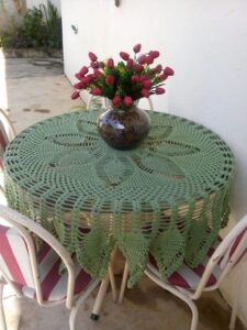 Toalha redonda de crochê verde sobre mesa com vaso de flores vermelhas, em ambiente doméstico externo