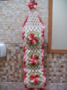 Porta papel higiênico de crochê pendurado na parede com flores em vermelho, branco e verde.