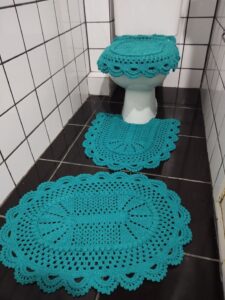 Jogo de banheiro em crochê azul com três peças sobre piso escuro e azulejos brancos.