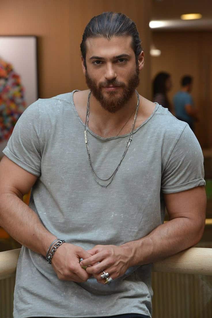 Ator turco Can Yaman com expressão séria, usando camiseta cinza e acessórios prateados, posando em ambiente interno.