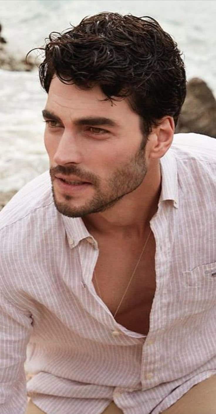 Ator turco Akin Akinözü com expressão pensativa, vestindo camisa clara, em ambiente externo.