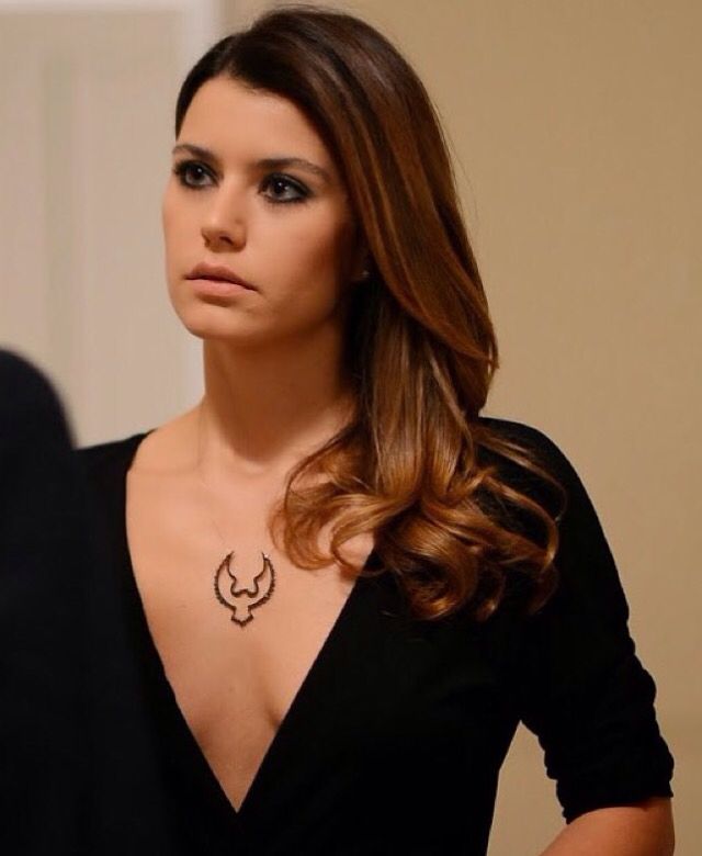 Atriz turca Beren Saat com expressão séria, usando vestido preto decotado e colar marcante, em ambiente interno.