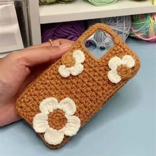  Capinha de celular em crochê marrom com flores brancas aplicadas, mostrando detalhes personalizados e acabamento artesanal.