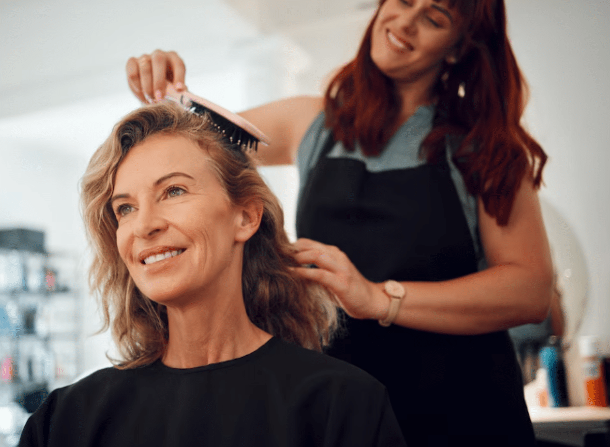 Mulher madura sorrindo enquanto tem o cabelo escovado por cabeleireira em salão de beleza