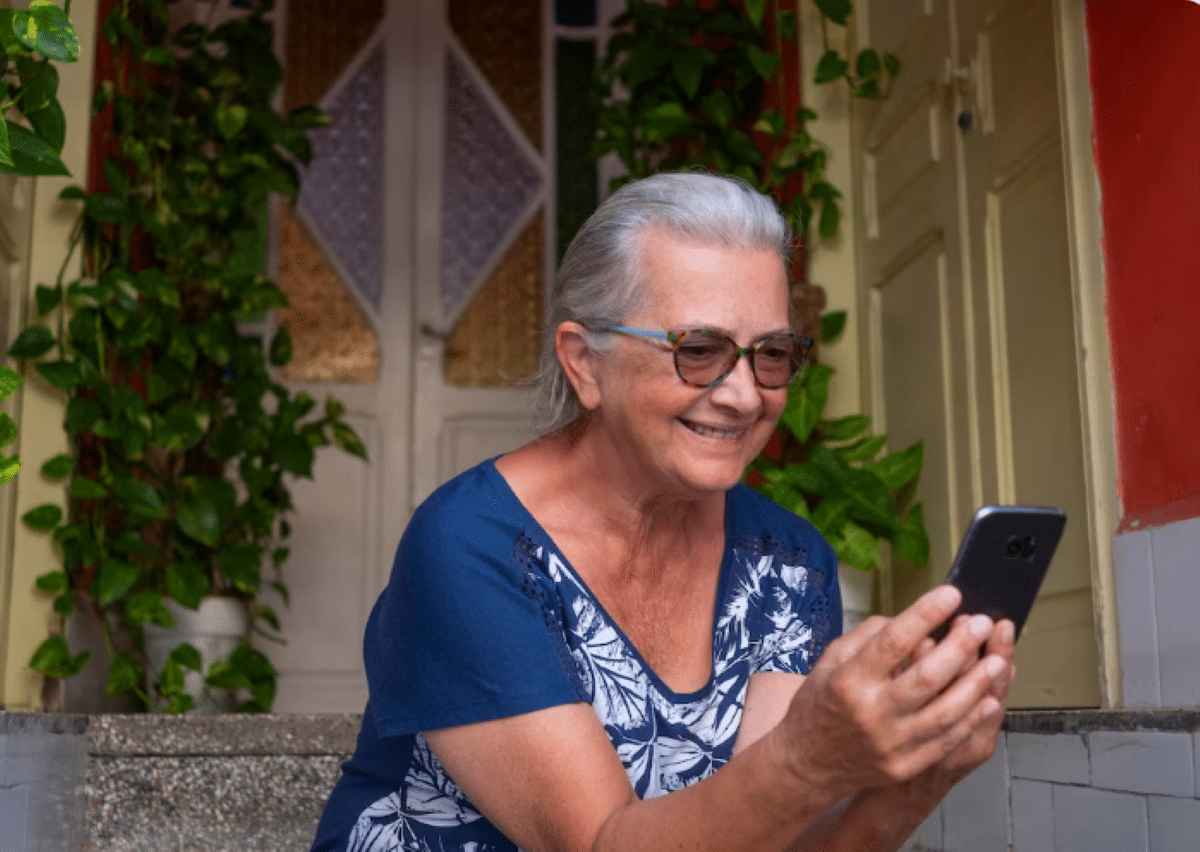 Mulher sorridente usando o celular sentada em frente à sua casa.