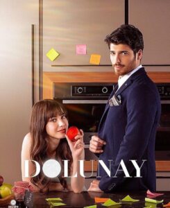 Casal da novela turca "Dolunay" em um momento descontraído na cozinha, com a mulher segurando uma maçã e o homem ao seu lado.