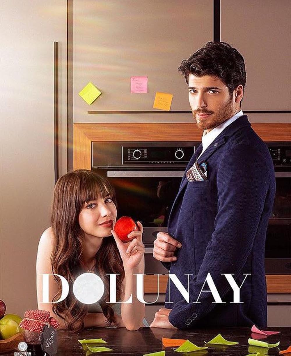 Casal da novela turca "Dolunay" em um momento descontraído na cozinha, com a mulher segurando uma maçã e o homem ao seu lado.